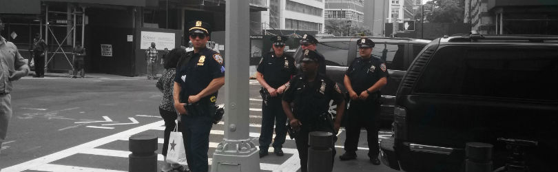 Image description: Five New York Police Department police officers in uniform are standing on the street with their hands on their belt. The officers are in color and the background is black and white.
