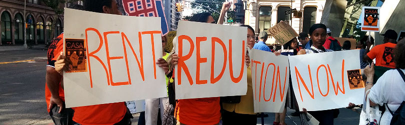 Image description: Four people are each holding a poster to form the words "RENT REDUCTION NOW." The words are in all caps and in orange. On the ends of the posters is an orange sticker with the words CASA and a fist.