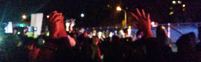 Image description: A grainy photo of two people's hands that are up in the air. One hand is holding onto another. The third hand is thrown up in the air in a "don't shoot" pose. There are protestors in the background.